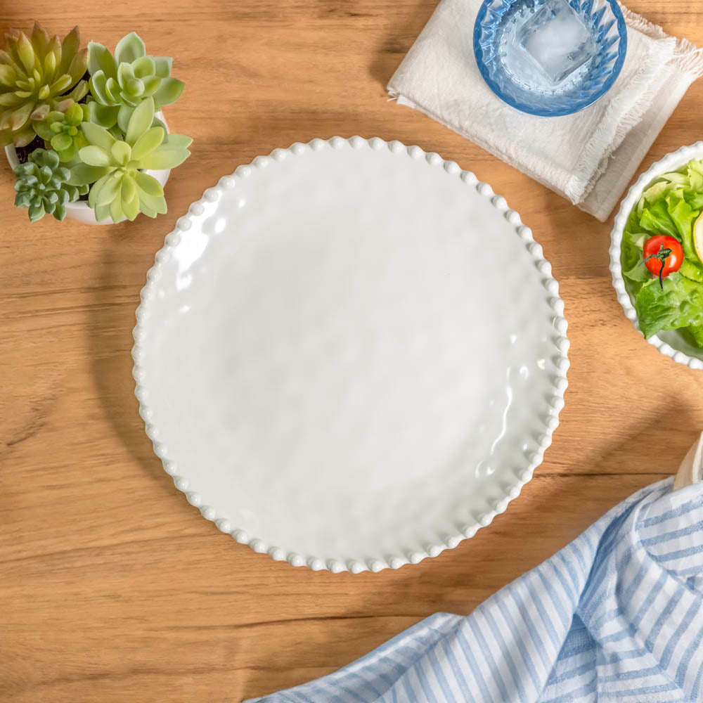 Cream colored, 11-inch melamine dinner plate set with beaded rim on a wooden table with acrylic tumbler, salad bowl, and napkin